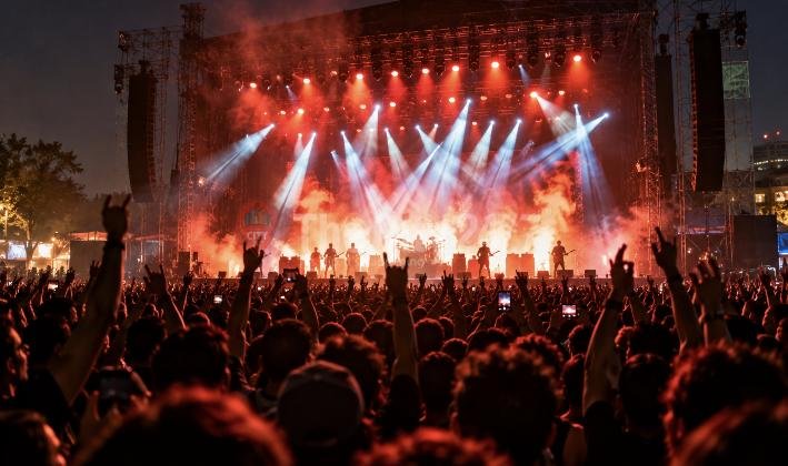 A rock band plays on a smoky outdoor stage lit by red and white spotlights as a huge crowd raises fists and horns