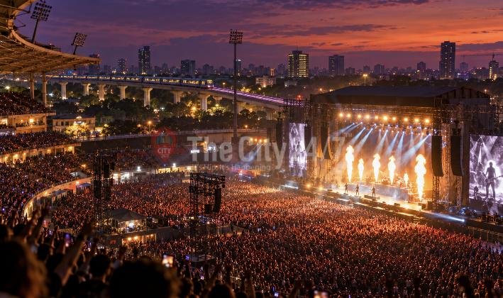 A huge crowd fills a stadium for a rock concert with flame jets and big screens on stage, a metro line and city skyline at dusk